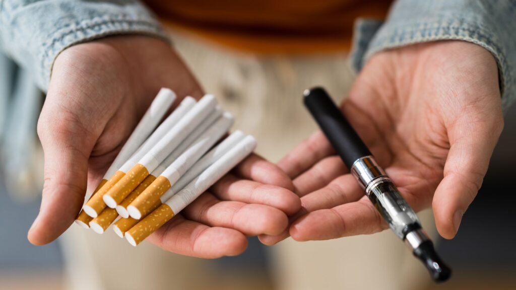 A person holds several traditional cigarettes in one hand and a vape pen in the other, illustrating a choice between smoking methods.