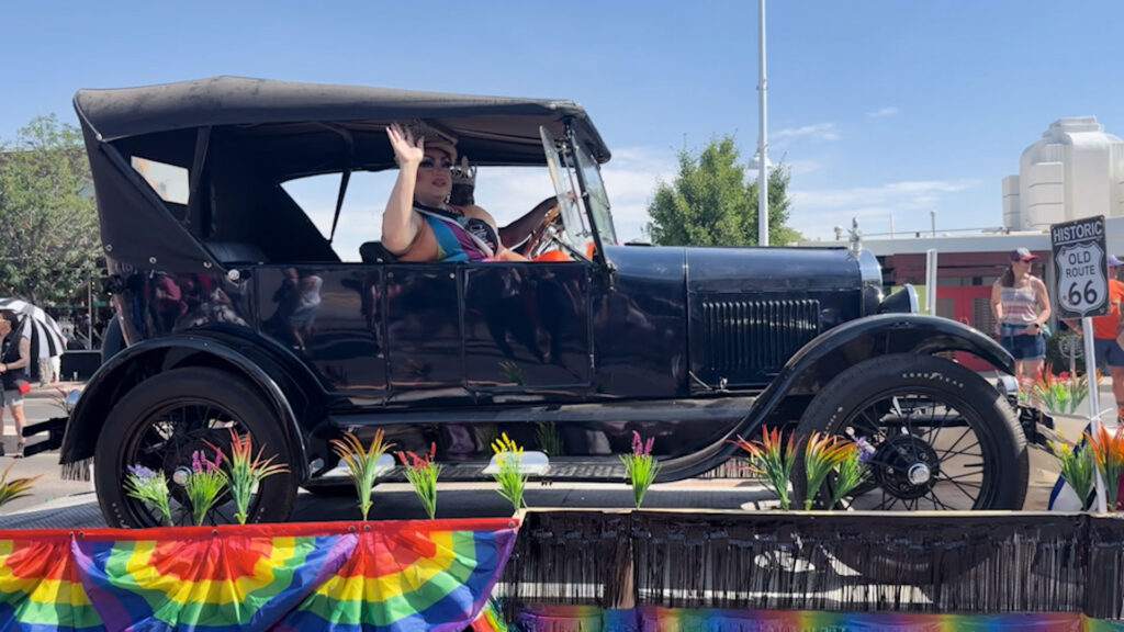 Person waves from a vintage black convertible decorated with rainbow flags during a parade, with a Historic Route 66 sign visible in the background.