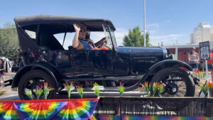 Person waves from a vintage black convertible decorated with rainbow flags during a parade, with a Historic Route 66 sign visible in the background.