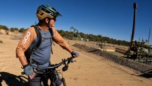 A cyclist wearing a helmet and gloves pauses on a dirt trail, looking toward oil pump jacks and equipment in a fenced area under a clear blue sky.