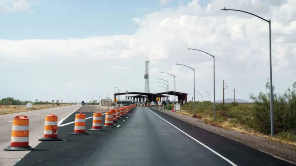 A highway checkpoint with orange traffic barrels narrowing lanes, leading to a covered inspection area under a cloudy sky.