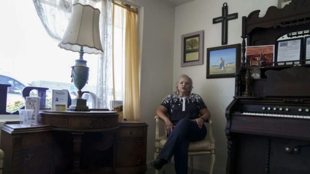 Woman sitting on a chair in a living room next to a lamp, piano, and wall with framed pictures and a cross.