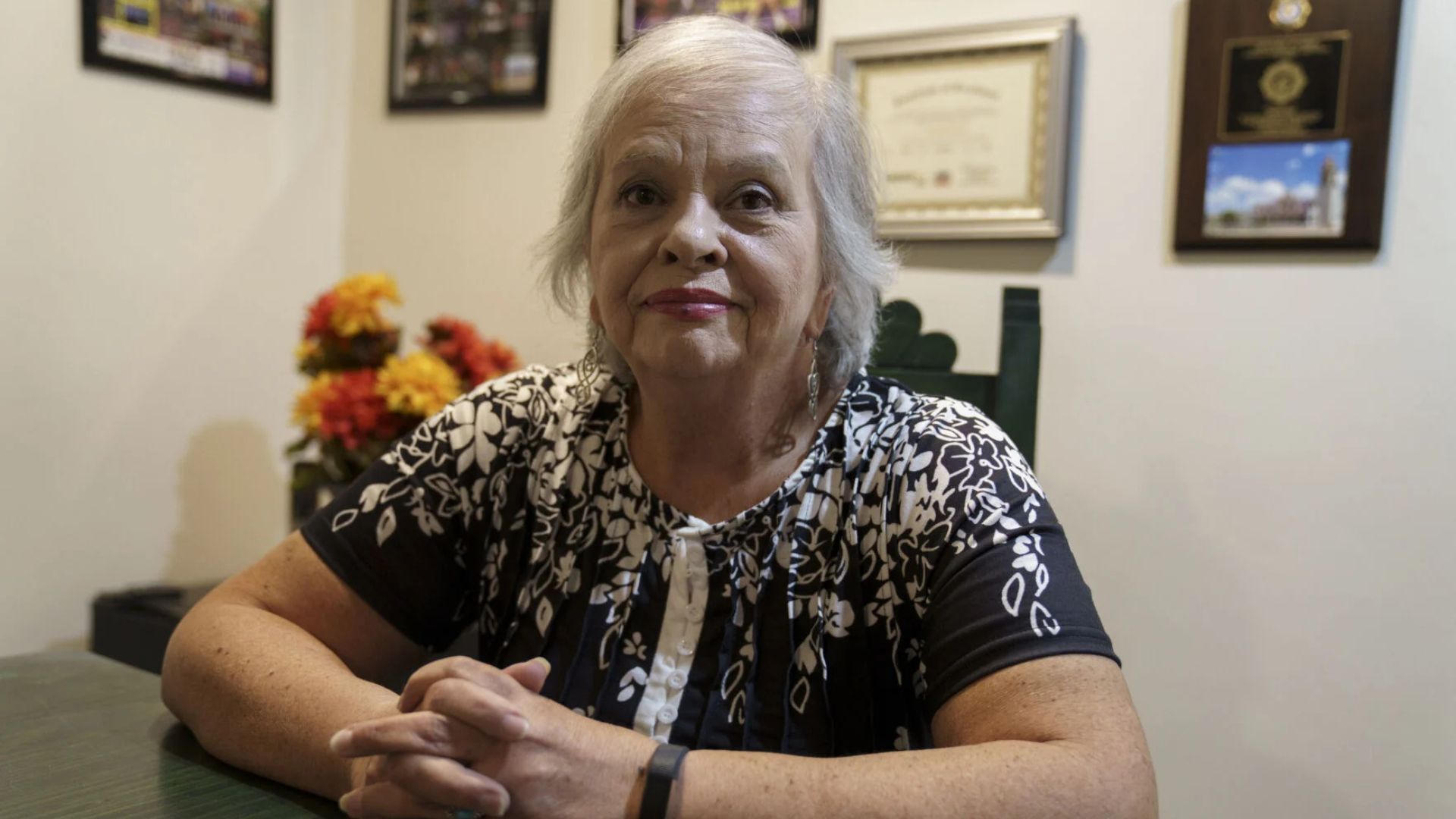 An older woman with gray hair sits at a table with her hands clasped, in a room decorated with framed certificates and flowers.