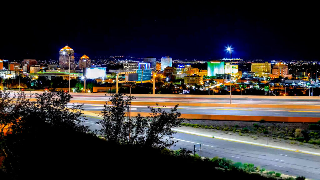 Night view of a city skyline with illuminated buildings and blurred car lights on a highway in the foreground. Trees line the edge of the road.