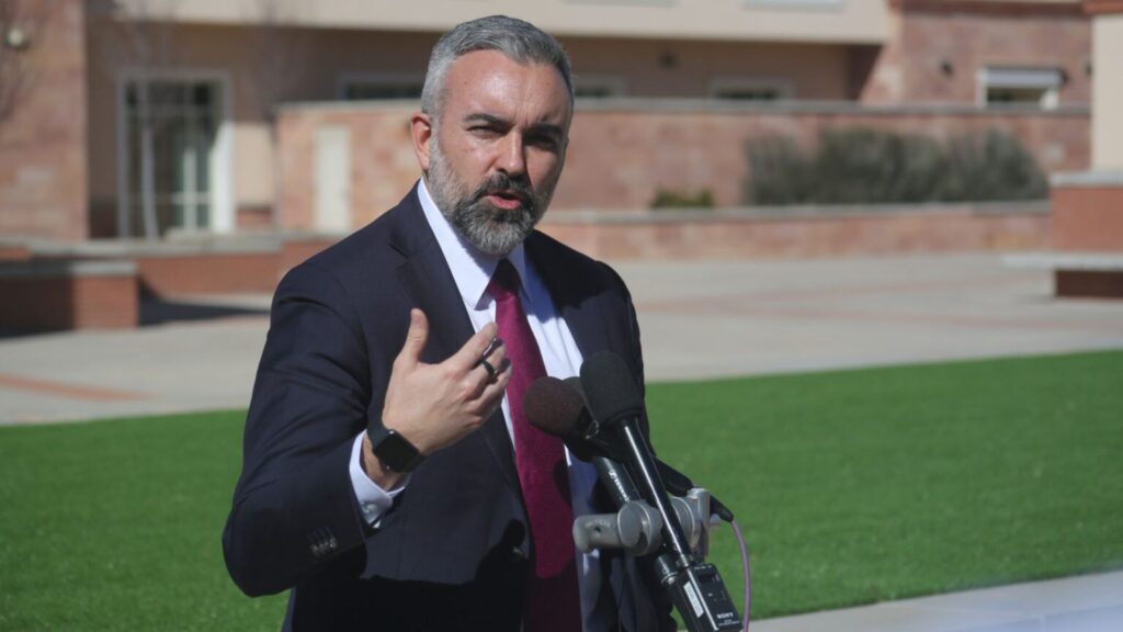 A man in a suit and tie speaks outdoors at a podium with microphones, gesturing with his hand. A building and green lawn are in the background.