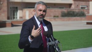 A man in a suit and tie speaks outdoors at a podium with microphones, gesturing with his hand. A building and green lawn are in the background.
