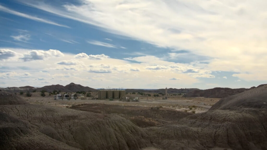 A dry, rocky desert landscape with scattered buildings and storage tanks under a partly cloudy sky.