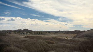 A dry, rocky desert landscape with scattered buildings and storage tanks under a partly cloudy sky.