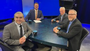 Four men in suits sit around a round table with blue mugs and papers, on a television studio set with blue background screens.