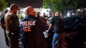 Several ICE officers in marked jackets gather around a dark vehicle at night, with blurred faces and trees visible in the background.