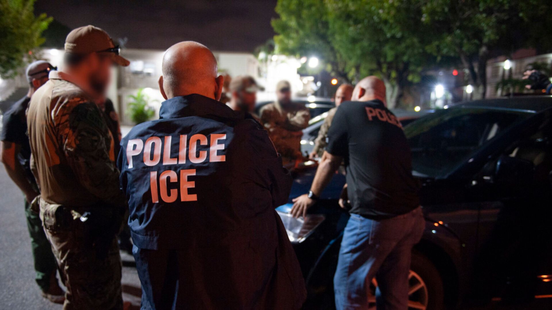 Several ICE officers in marked jackets gather around a dark vehicle at night, with blurred faces and trees visible in the background.
