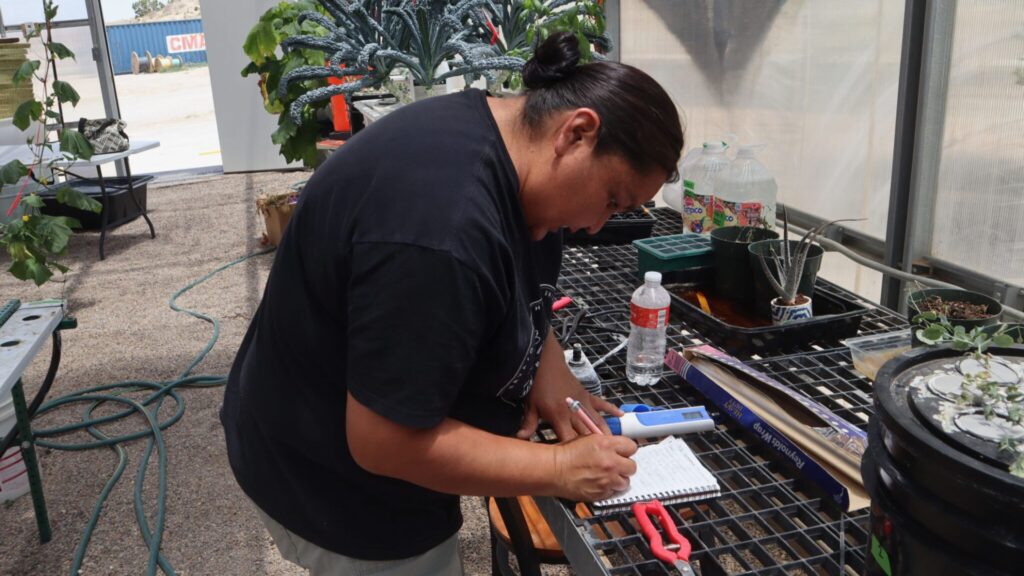 Person writing in a notebook while standing at a greenhouse table with gardening tools, plants, and water bottles nearby.