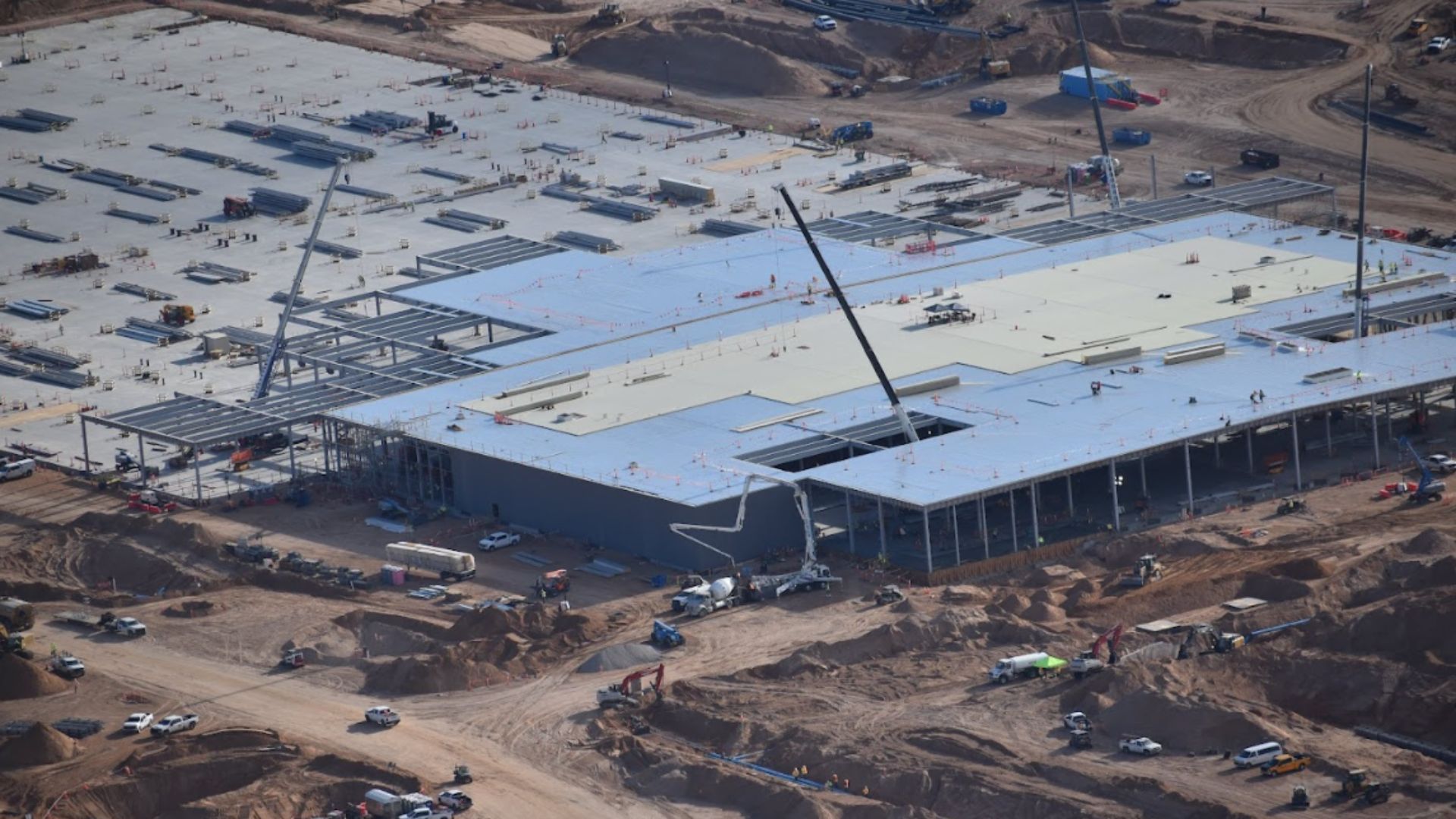 Aerial view of a large construction site with steel framework, cranes, construction vehicles, and workers assembling a building surrounded by dirt and equipment.