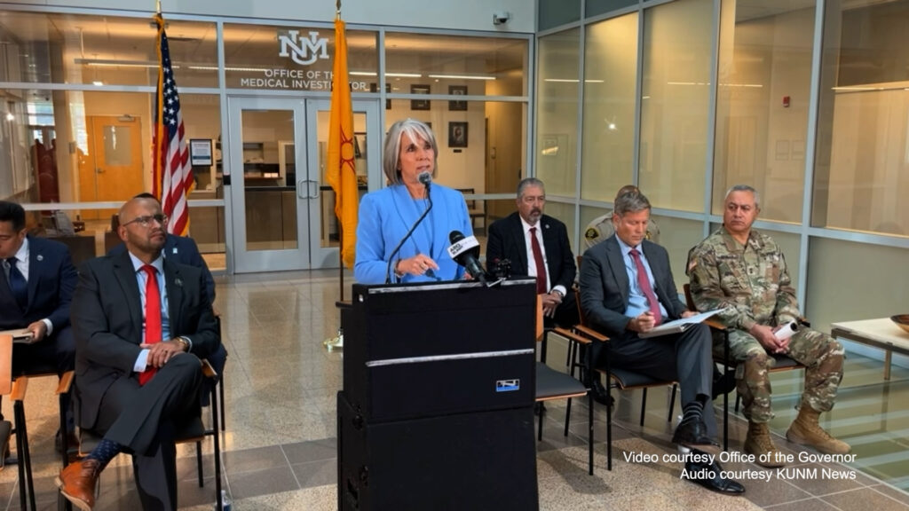 New Mexico governor speaks at a podium in an indoor setting, flanked by officials including law enforcement and military personnel.