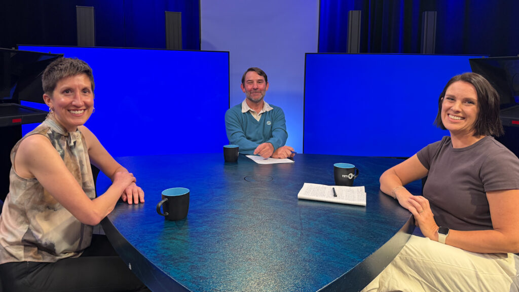 Three people sit at a round table with blue backgrounds, mugs, and a notepad in a studio setting, facing the camera and smiling.