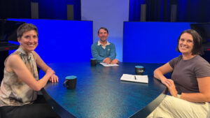 Three people sit at a round table with blue backgrounds, mugs, and a notepad in a studio setting, facing the camera and smiling.
