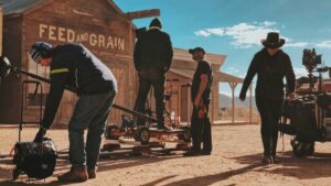 Film crew sets up equipment on a dusty outdoor set with a wooden “Feed and Grain” building in the background under a sunny sky.