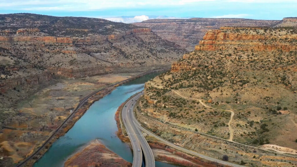 A winding highway runs alongside a river through a desert canyon with layered rock formations and sparse vegetation.