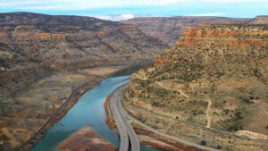 A winding highway runs alongside a river through a desert canyon with layered rock formations and sparse vegetation.