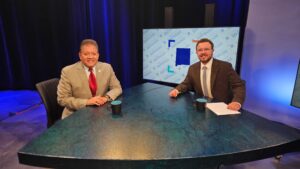 Two men in suits sit at a triangular news desk with two mugs and papers, facing the camera; a screen in the background displays the words “in focus.”.