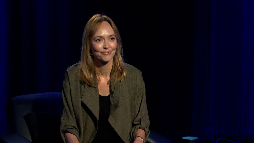 A woman with light brown hair wearing a dark jacket sits on a stage in front of a dark background, facing the camera and smiling slightly.
