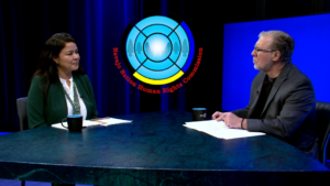 Two people sit at a table having a discussion in a studio with a "Navajo Nation Human Rights Commission" logo displayed on a screen behind them.