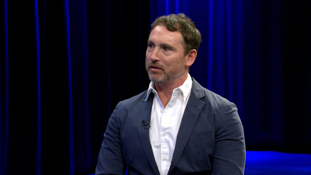 A man with short brown hair and a beard, wearing a white shirt and navy blazer, sits and talks in front of a dark blue curtain background.