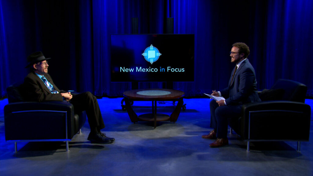 Two men in suits sit across from each other in a TV studio with blue lighting. A screen behind them displays the logo and text "New Mexico in Focus.