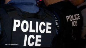 Two law enforcement officers are shown from the back, wearing jackets labeled “POLICE ICE,” standing close together in a dimly lit environment.