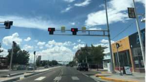 A city street with cars stopped at a red traffic light under a partly cloudy sky, with shops and trees lining both sides of the road.