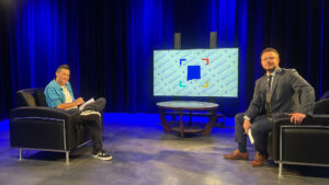 Two men sit in armchairs on a studio set with a round table and a large screen displaying “In Focus” in the background. One man wears casual clothes, the other wears a suit.