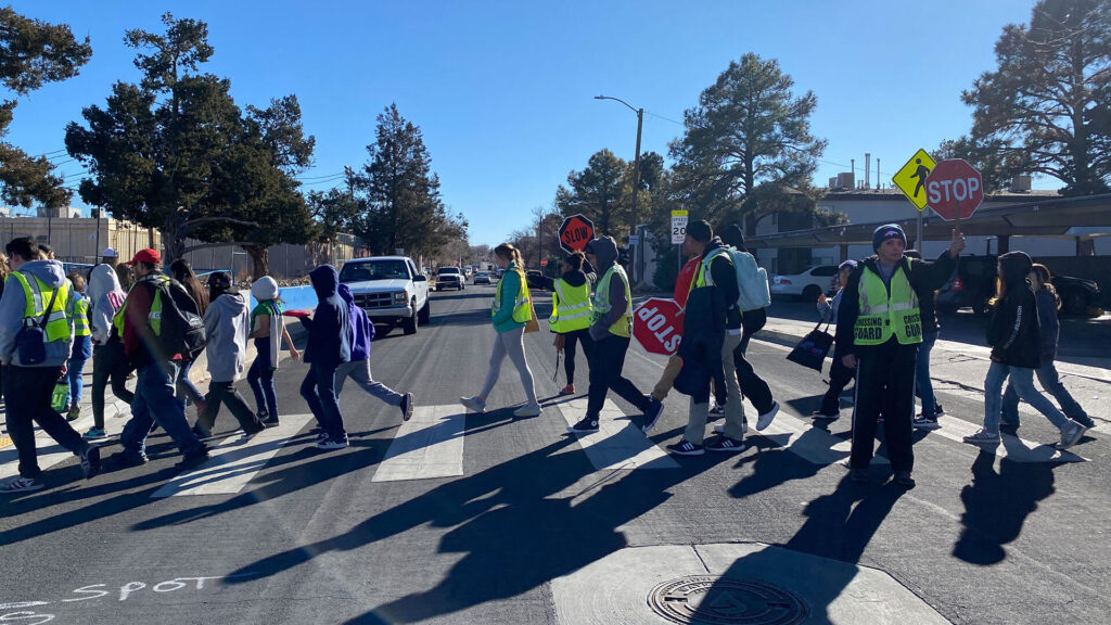 A group of people, some wearing safety vests and holding stop signs, guide children and adults across a crosswalk on a sunny day.
