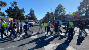A group of people, some wearing safety vests and holding stop signs, guide children and adults across a crosswalk on a sunny day.
