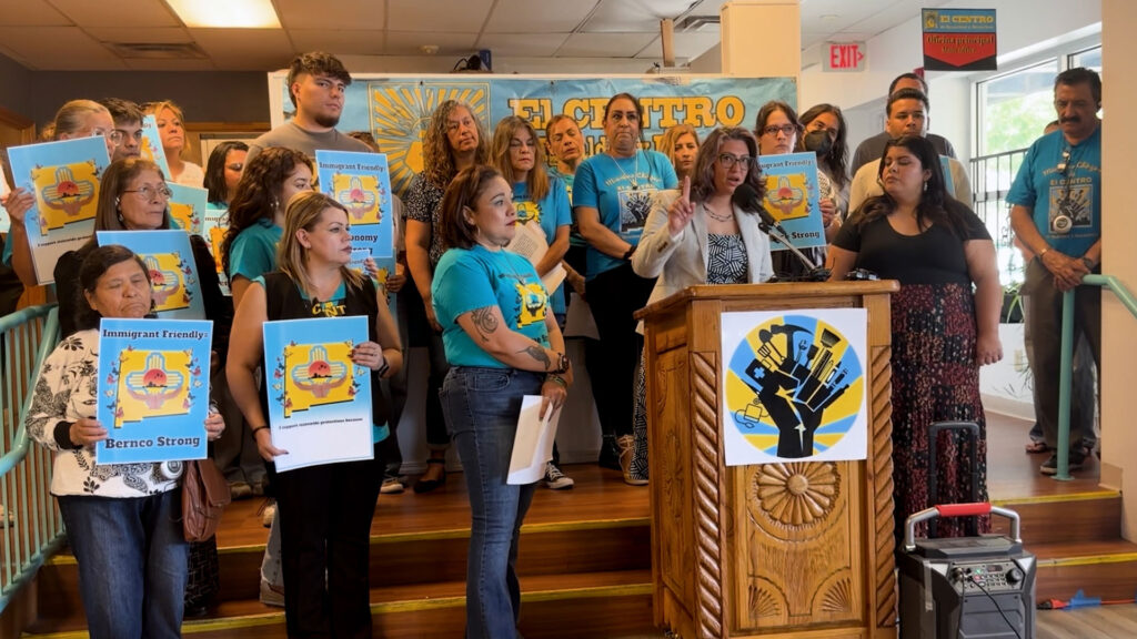 A group of people stands on steps as a woman speaks at a podium. Many hold signs with “Immigrant Friendly” messages. A banner behind them reads “El Centro.”.