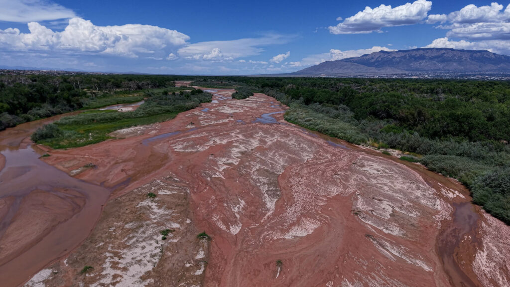 A mostly dry riverbed with patches of water winds through a green landscape under a blue sky with clouds and distant mountains.