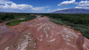A mostly dry riverbed with patches of water winds through a green landscape under a blue sky with clouds and distant mountains.