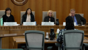 A person sits at a desk facing four officials in a wood-paneled meeting room, with nameplates and microphones visible on the officials’ table.