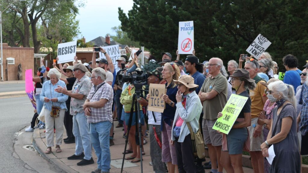 A group of people stand on a sidewalk holding signs and banners at a protest or rally, with trees and buildings in the background.