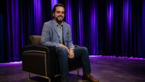 A man in a blazer sits on a black chair in front of purple-lit stage curtains, smiling at the camera.
