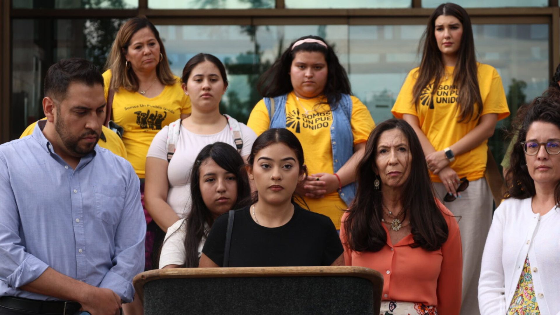 A group of people stands behind a podium outdoors; some wear yellow shirts with a logo, while others wear casual clothing, looking serious.