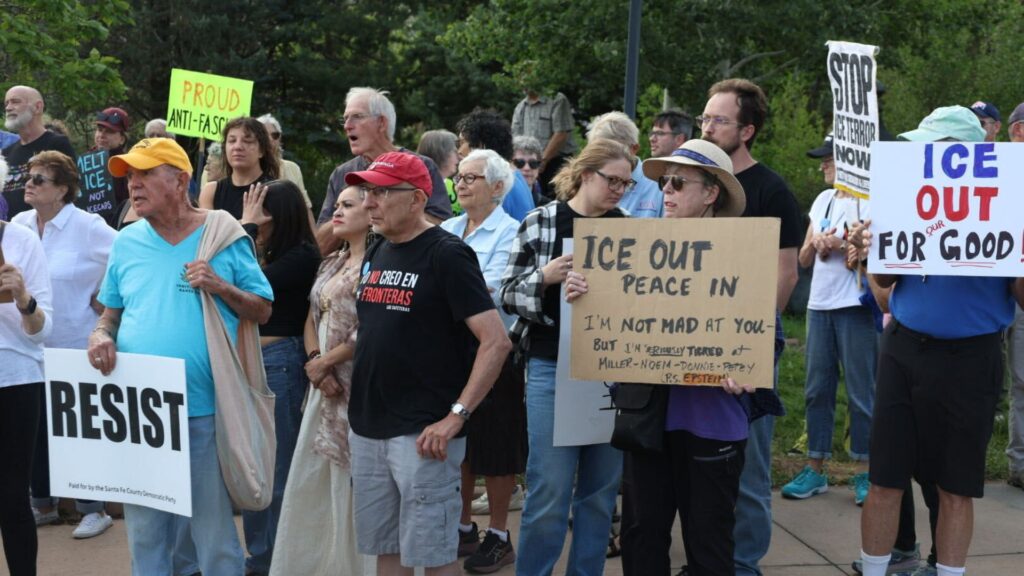 A group of people stand outdoors holding protest signs with messages such as “RESIST,” “ICE OUT,” and “STOP ICE NOW.” Some people are talking while others face forward.