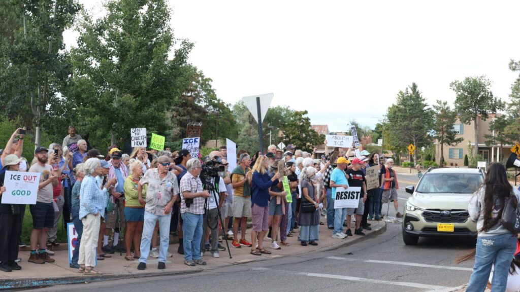 A group of people stand on a street corner holding protest signs, including messages such as “RESIST” and “ICE OUT,” while some interact with a person by a parked car.