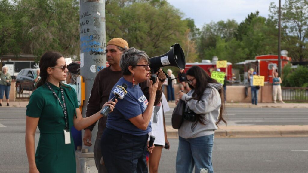 A woman speaks into a megaphone while another holds a microphone labeled “13,” with several people and protest signs visible in the background.