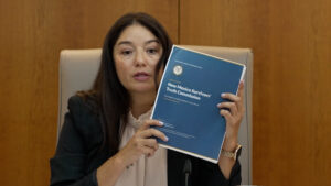 A woman sits at a desk holding up a blue report titled “New Mexico Survivors’ Truth Commission” in front of a wooden background.