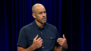 A bald man in a buttoned short-sleeve shirt gestures with his hands while speaking in front of dark blue curtains.