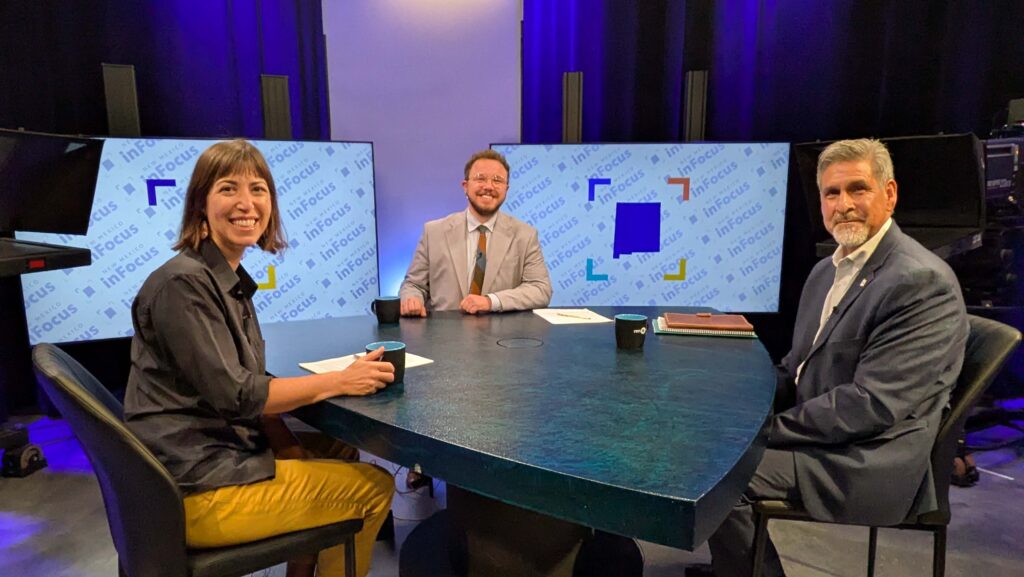 Three people sit around a round table in a TV studio with two “In Focus” screens in the background, appearing ready for a discussion.