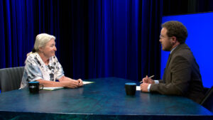 Two people sit across from each other at a table in a studio, having a conversation. One holds a pen and notepad; the set is lit with blue lighting.