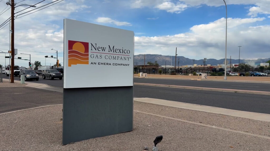 A New Mexico Gas Company sign stands by a road with cars, businesses, and mountains visible in the background under a partly cloudy sky.