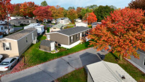Aerial view of a mobile home park in autumn, with colorful trees, several mobile homes, green grass, and paved roads.