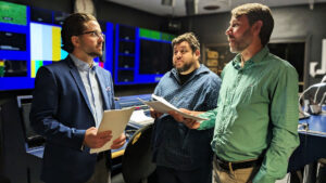 Three men stand in a control room, holding papers and having a discussion. Monitors and technical equipment are visible in the background.